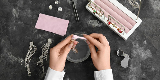 On a hard surface, some silver pieces and a pink cloth lay while a person's hands brush on a silver jewelry.